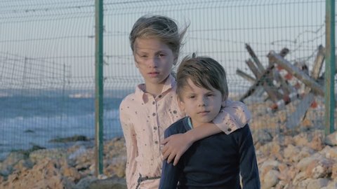 orphan Depressed children. Loneliness girl crying near to sad boy. Sad little refugees Brother and sister on the shore near the state border. protected area with barbed wire fence by the sea