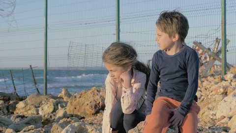 two poor refugee children boy girl sitting on the stones near the sea. fence barbed wire separating the state. concept sadness and suffering