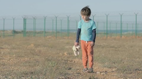 lonely sad boy orphan walking along the abandoned road with head down in the hands of holding a toy rabbit stopping and looking into the distance. concept of immigration and refugee camp