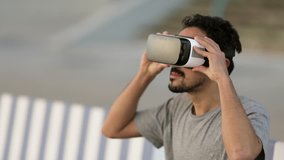 Young Arabic man with dark curly hair and beard in grey T-shirt sitting in park on striped bench in virtual reality glasses, investigating new reality. High technology, VR concept