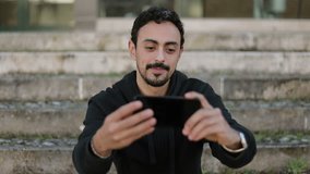 Front view of young Arabic man with dark curly hair and beard in black hoodie sitting on stairs outside, having video chat with friend, waving hand. Lifestyle, communication concept