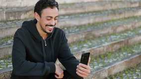 Young Arabic man with dark curly hair and beard in black hoodie sitting on stairs outside, having video chat, waving hand, showing surroundings. Lifestyle, communication concept
