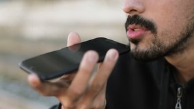 Close up shot of young Arabic mans face with dark curly hair and beard in black hoodie sitting on stairs outside, talking on phone with speakerphone. Lifestyle, communication concept