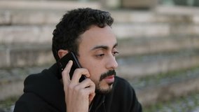 Closeup of young Arabic mans face with dark curly hair and beard in black hoodie sitting on stairs outside, talking on phone, drinking takeaway coffee. Lifestyle, communication concept