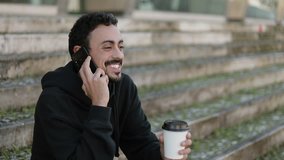 Young Arabic man with dark curly hair and beard in black hoodie sitting on stairs outside, talking on phone, smiling and holding takeaway coffee in hand. Medium shot. Lifestyle, communication concept