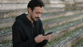 Young Arabic man with dark curly hair and beard in black hoodie sitting on stairs outside, holding phone in hands, texting and smiling. Medium shot. Lifestyle, communication concept