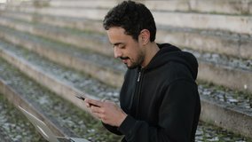 Young Arabic man with dark curly hair and beard in black hoodie sitting on stairs outside, holding phone in hands, texting, smiling, holding laptop on knees. Lifestyle, communication concept