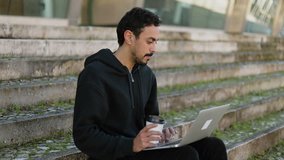 Young Arabic handsome man with dark curly hair and beard in black hoodie sitting on stairs outside, working on laptop, drinking takeaway coffee. Lifestyle, work concept