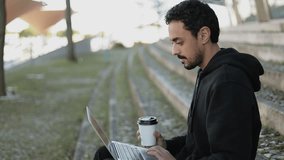 Side view of young Arabic handsome man with dark curly hair and beard in black hoodie sitting on stairs outside, working on laptop, drinking takeaway coffee. Lifestyle, work concept