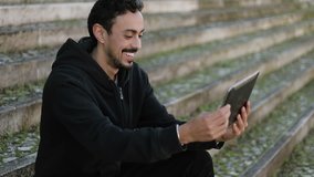 Side view of excited young Arabic handsome man with dark curly hair and beard in black hoodie sitting on stairs outside, swiping photos on tablet. Lifestyle, leisure concept