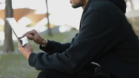 Medium shot of young Arabic handsome man with dark curly hair and beard in black hoodie sitting on stairs outside, swiping, pinching photos on tablet. City background. Lifestyle, leisure concept