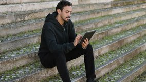 Young Arabic handsome man with dark curly hair and beard in black hoodie sitting on stairs outside, swiping photos on tablet. City background. Dolly shot. Lifestyle, leisure concept