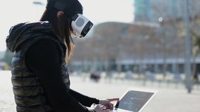 Side view of young dark-haired woman in warm black waistcoat sitting on stairs outside, working on laptop looking through virtual reality glasses. City background. VR concept