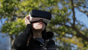 Young dark-haired woman in warm black waistcoat wearing virtual reality glasses in early autumn park, turning head, checking set up device parameters. High technology, VR concept