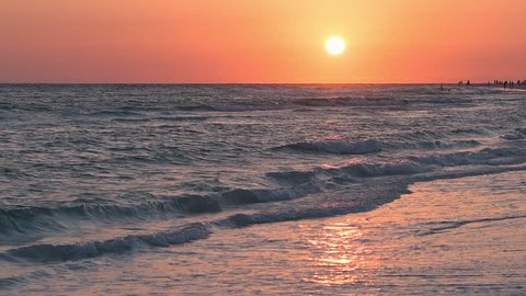 Slow motion of dreamy pink pastel orange sunset in Sarasota Siesta Key, Florida with  ocean coastline coast, waves crashing washing on beach shore, people silhouettes walking in far distance woman