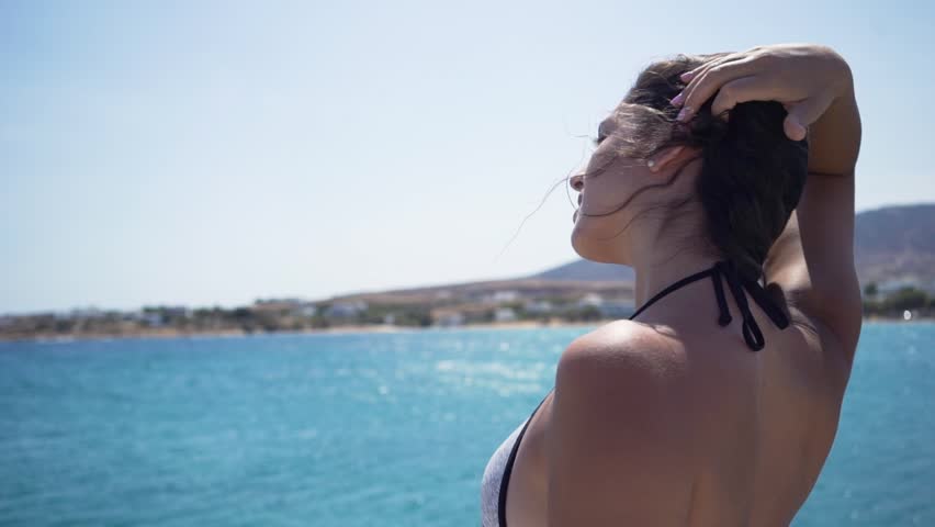 A young, beautiful woman looks out over the Aegean Sea in Paros, Greece; her hair blows in the wind; slow motion; static shot.