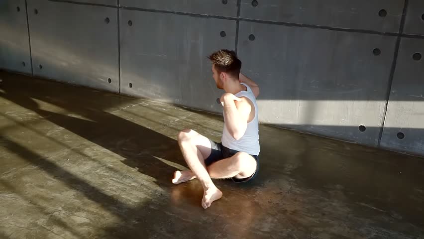 male athlete is making twists of torso sitting on floor in gymnastic room, warming shoulder girdle