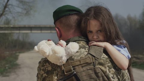 A Soldier Is Taking Out A Little Girl From A Combat Zone. A Man Carries A Child In His Arms. Crying, Frightened Child. Ruined City.