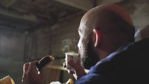 a guy with a bald head and a beard is drinking a hot drink from a disposable cup and biting a bun