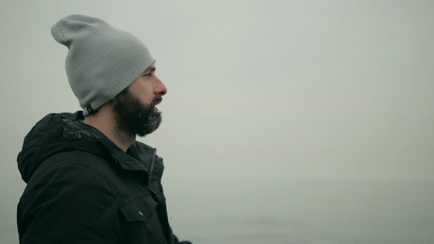 bearded man in a black jacket and a gray hat on the beach shows off. slow motion