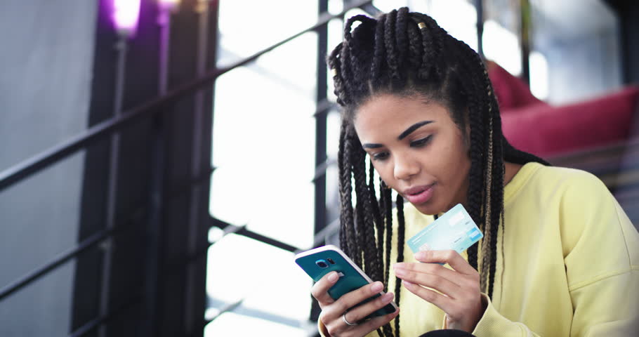 Charismatic african lady closeup buy something online using a phone and a bank card she sitting on the stairs in a modern building office. 4k
