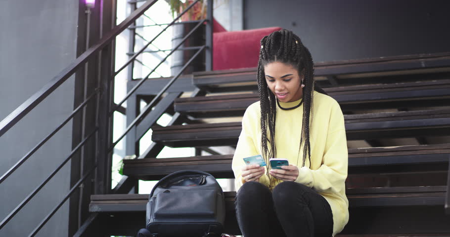 Good looking young african lady using her smartphone to order something online from her bank card. she happy at the end lifted the hands up, she sitting down on the modern stairs.