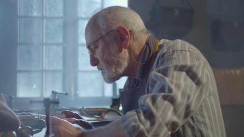 Footage of goldsmith working at table in workshop . Bearded old man jeweler creating new jewelry . Master goldsmith working process . Scenic film shot in vintage style. Shot on RED EPIC Camera.   
