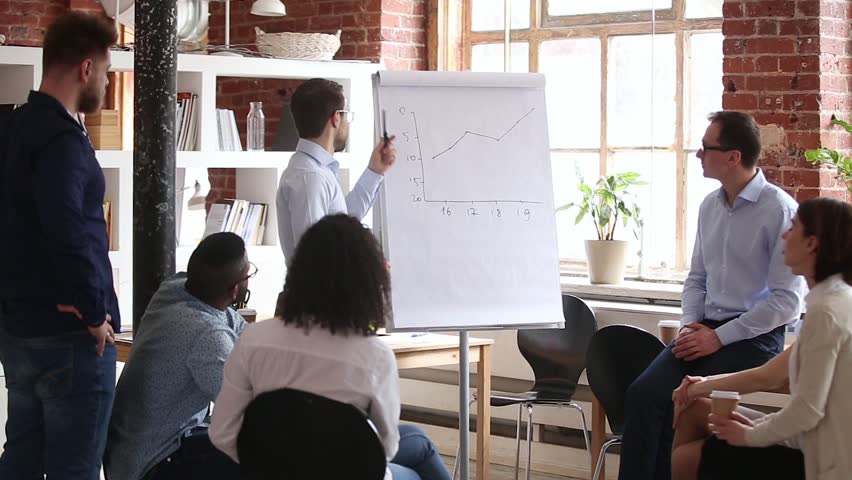 Confident speaker coach gives corporate presentation on whiteboard teaching diverse sales team at group meeting, male business trainer presenter speaking at training explaining report graph in office Royalty-Free Stock Footage #1024307081