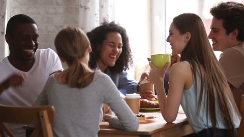Diverse happy students talking joking having fun share cafe table at group meeting, african and caucasian young friends laughing drinking coffee tea together enjoy multiracial friendship in dormitory
