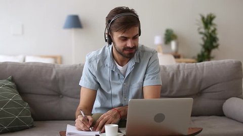 Happy young man in headset speaking by webcam looking at laptop make notes, male sales representative talking by video call study online consulting customer selling service support working from home