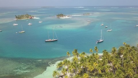 A beautiful drone shot of some sailboats anchoring in a blue lagoon between a few caribbean islands overgrown with green palm trees on a sunny day in San Blas, Panama.