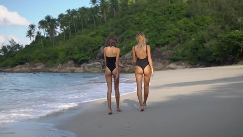 two long-legged models in black bikini are on the ocean beach, going at the edge of ocean water gilded by the level rays of the sun