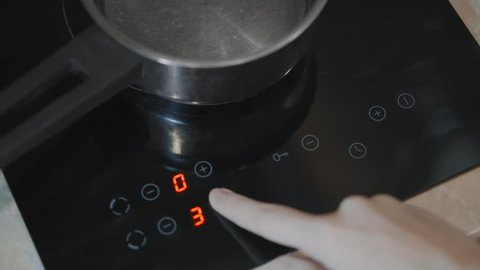 A close-up of a female hand includes an electric stove. Modern technology. Touch panel of electric cooker. Metal pan stands on the touch plate.