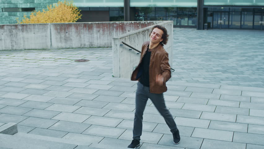 Happy Young Man with Long Hair Actively Dancing While Walking Up the Stairs. He's Wearing a Brown Leather Jacket. Scene Shot in an Urban Concrete Park Next to Business Center. | Shutterstock HD Video #1024342472