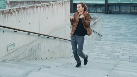 Happy Young Man with Long Hair Actively Dancing While Walking Up the Stairs. He's Wearing a Brown Leather Jacket. Scene Shot in an Urban Concrete Park Next to Business Center.