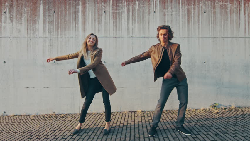 Cheerful Girl and Happy Young Man with Long Hair are Actively Dancing Meme Moves on a Street next to an Urban Concrete Wall. They Wear Brown Leather Jacket and Coat. Sunny Day. Royalty-Free Stock Footage #1024342517