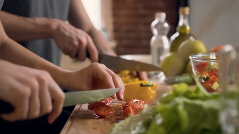 Close up shot of man hands slicing carrot and female hands cutting tomato on wooden cutting board for salad on the table with healthy food in the kitchen. Side view.