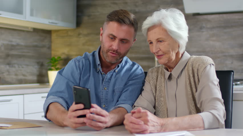 Lockdown of adult man sitting with his senior mother at kitchen table, holding smartphone in his hands and making selfie