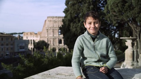 Portrait of a smiling Italian boy sitting on a stone ledge in a park with trees and the Coliseum in the background on a sunny day in Rome. Wide to medium shot on 4k RED camera.