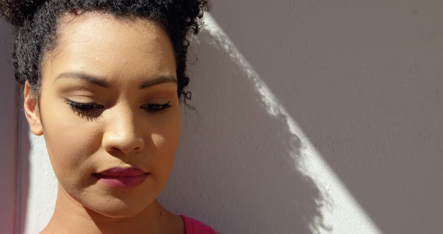 Front view close up of young mixed-race woman leaning against the wall in backyard of home. She is looking down 4k