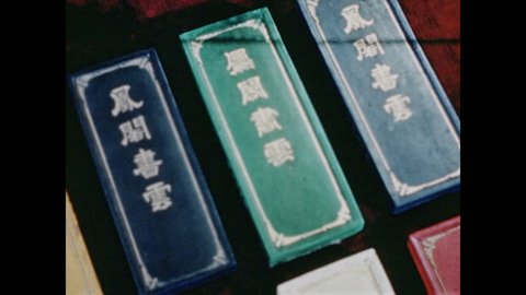 1940s: UNITED STATES green and blue rectangular boxes with Chinese labels sit on floor. hand picks up red box. fingers drip water from kettle onto white paper circle and rubs block to create watercolor paint.