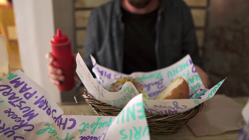 Guy is seasoning hamburger by tomato ketchup. He is pressing plastic bottle, sitting in fast food cafe, close-up view