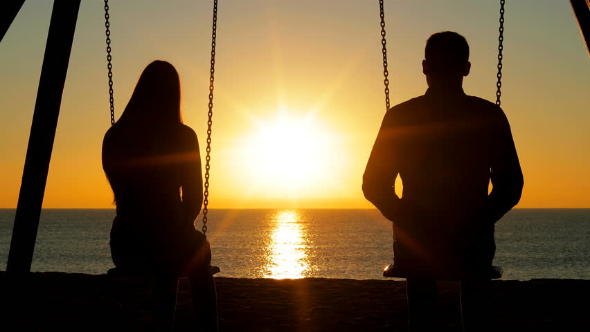 Back view silhouette of a happy couple swinging holding hands at sunrise on the beach