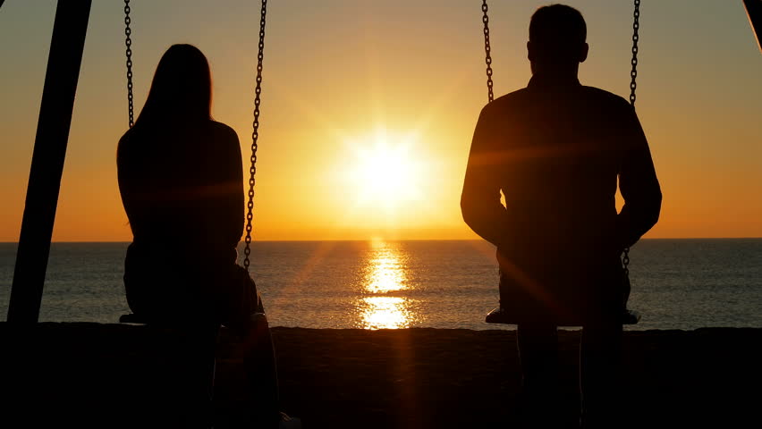 Back view silhouette of a happy couple swinging looking each other at sunrise