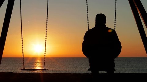 Back view silhouette of a sad man alone swinging looking at empty seat at sunrise on the beach