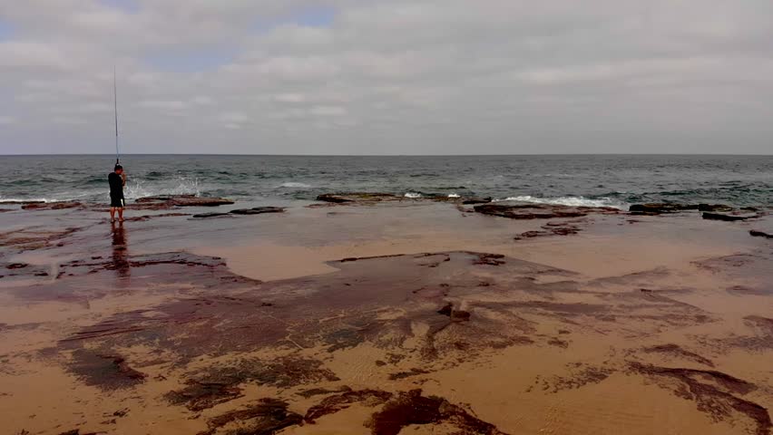 Aerial footage of man rock fishing with large waves crashing on rocks near by.