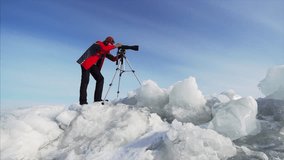 Sliding view from below of photographer with camera on huge ice blocks taking photos or video of frozen lake. Blue sky is on the background