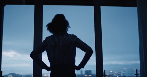 young woman looking out window enjoying view of city at night contemplating successful lifestyle planning ahead on calm urban evening