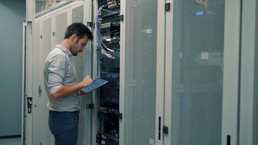 Male It Technician Engineer With Tablet Work In Server Room In Data Center.  Male IT Engineer Working In A Data Center With Rows Of Server Racks And Super Computers, He Is Looking Into Data Cabinets Royalty-Free Stock Footage #1024448702