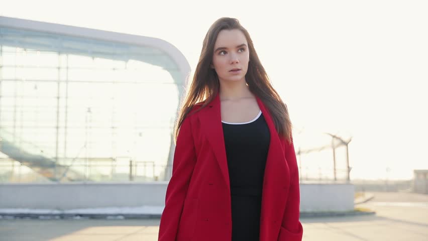 beautiful business woman in red coat goes near business centre in the city, communicates via smartphone (phablet). Steady cam shot, slow motion.autumn look
